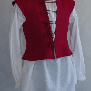 Puede incluir: Un chaleco rojo oscuro sin mangas con cuello alto y cierre frontal mediante cordones, usado sobre una camisa blanca de manga larga con escote sencillo y pantalones verdes, exhibido en un maniqu&iacute; sobre un fondo de color neutro.