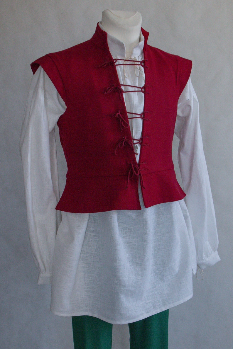 Puede incluir: Un chaleco rojo oscuro sin mangas con cuello alto y cierre frontal mediante cordones, usado sobre una camisa blanca de manga larga con escote sencillo y pantalones verdes, exhibido en un maniqu&iacute; sobre un fondo de color neutro.