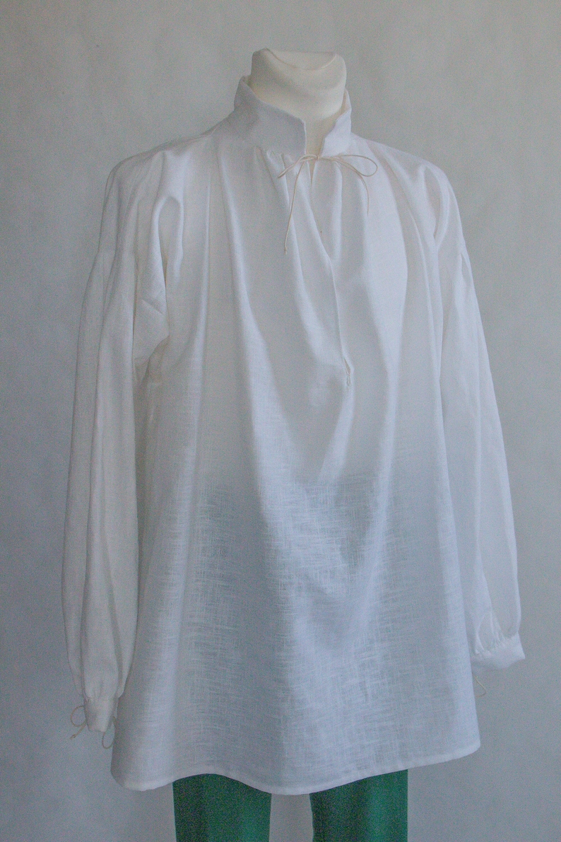 Puede incluir: Una camisa blanca de manga larga con cuello alto y un fino cord&oacute;n ajustable en el escote. La prenda presenta mangas amplias y fruncidas que se estrechan en las mu&ntilde;ecas con cordones a juego, creando una silueta holgada y fluida.
