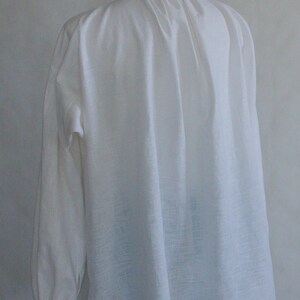Puede incluir: Una camisa blanca de manga larga con cuello alto y tela fruncida en el escote, creando pliegues verticales en la espalda. La prenda tiene mangas amplias con pu&ntilde;os y presenta un tejido de textura visible en toda su superficie.