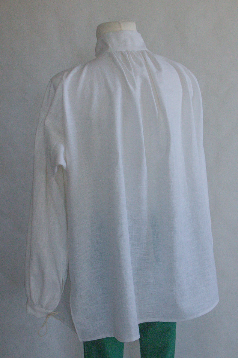Puede incluir: Una camisa blanca de manga larga con cuello alto y tela fruncida en el escote, creando pliegues verticales en la espalda. La prenda tiene mangas amplias con pu&ntilde;os y presenta un tejido de textura visible en toda su superficie.