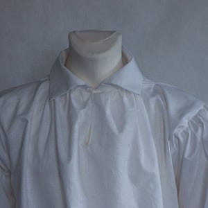 May include: A long-sleeved white shirt featuring a classic fold-down collar and gathered fabric detailing at the shoulders. The garment is displayed on a mannequin, showcasing a plain, solid white textile construction with a button-down front placket.