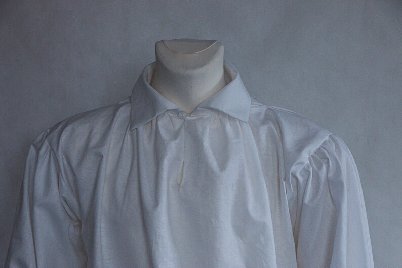 May include: A long-sleeved white shirt featuring a classic fold-down collar and gathered fabric detailing at the shoulders. The garment is displayed on a mannequin, showcasing a plain, solid white textile construction with a button-down front placket.