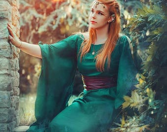 May include: A floor-length emerald green dress with wide, flowing sleeves and a dark maroon waist sash. The garment features a simple round neckline and a long, loose skirt that drapes down. The fabric appears lightweight and opaque in texture.