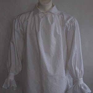 May include: A long-sleeved white shirt featuring a classic collar, gathered fabric at the shoulders, and wide cuffs with flared ruffles at the wrists. The garment is displayed on a mannequin, showcasing a loose fit and a button-down front closure.