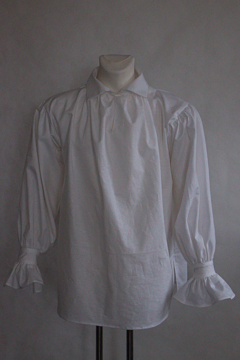 May include: A long-sleeved white shirt featuring a classic collar, gathered fabric at the shoulders, and wide cuffs with flared ruffles at the wrists. The garment is displayed on a mannequin, showcasing a loose fit and a button-down front closure.