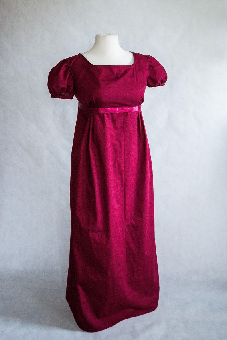 May include: A floor-length, deep magenta dress featuring a square neckline, short puffed sleeves with gathered detailing, and a high empire waistline accented by a thin, matching satin ribbon band that encircles the torso above the skirt.