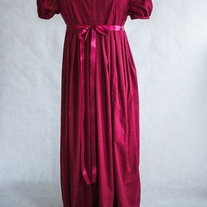 May include: A floor-length, deep magenta dress featuring short puff sleeves, a high empire waistline, and a matching satin ribbon tied in a bow at the center back. The garment has a wide, rounded neckline and a full, gathered skirt that falls straight.