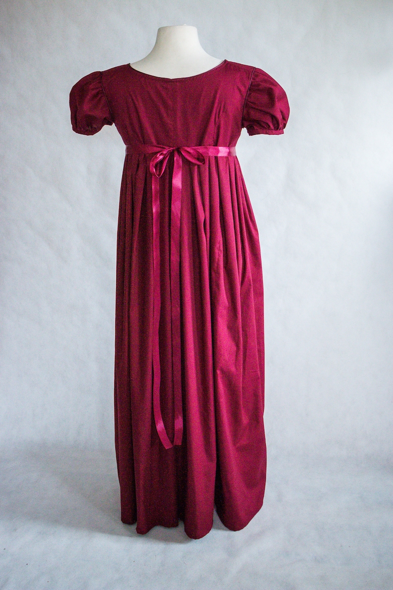 May include: A floor-length, deep magenta dress featuring short puff sleeves, a high empire waistline, and a matching satin ribbon tied in a bow at the center back. The garment has a wide, rounded neckline and a full, gathered skirt that falls straight.