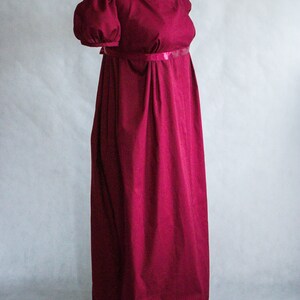 May include: A floor-length, deep burgundy dress featuring a high empire waistline, short puffed sleeves, and a square neckline. The garment is accented with a thin, matching satin ribbon band directly under the bust, with a long, flowing skirt.
