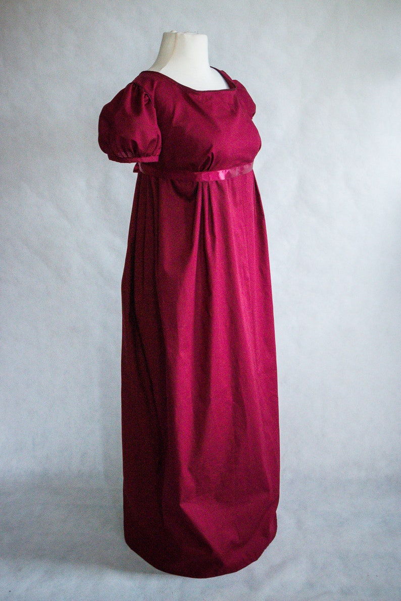 May include: A floor-length, deep burgundy dress featuring a high empire waistline, short puffed sleeves, and a square neckline. The garment is accented with a thin, matching satin ribbon band directly under the bust, with a long, flowing skirt.