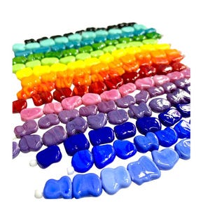 Könnte beinhalten: Eine Regenbogen-Auswahl an Glasperlen in verschiedenen Blautönen, Lila, Rosa, Rot, Orange, Gelb, Grün und Blau. Die Perlen haben alle die Form eines Tropfens und sind in einer Reihe angeordnet.