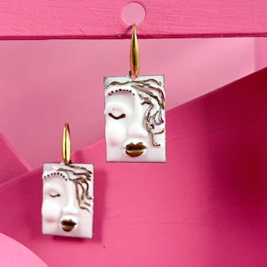 May include: A pair of rectangular drop earrings featuring a white enamel face design with gold-toned metallic accents highlighting the hair, closed eyes, and lips. Each earring hangs from a gold-toned metal hook, showcasing a stylised human face.