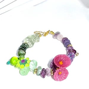 May include: A vibrant beaded bracelet with a mix of gemstones and decorative features. The bracelet showcases green, purple, and pink beads, complemented by gold-coloured details. The design incorporates floral and geometric shapes, creating a unique accessory.