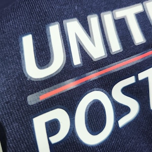 May include: A dark navy blue fabric garment featuring white block lettering that reads UNIT and POST. A thin horizontal red line is positioned between the two words. The text appears to be a screen-printed graphic on a textured cotton material.