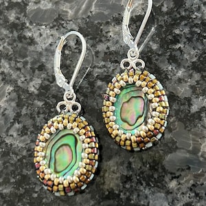 May include: A pair of dangle earrings featuring oval-shaped abalone shell cabochons surrounded by intricate beadwork in shades of metallic gold, bronze, and silver. The earrings are attached to silver-toned metal lever-back ear wires.