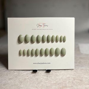 May include: A set of sage green press-on nails arranged on a white card. The card features the text "Sharp Talons Press On Nails" and the website address. The nails are oval-shaped and come in various sizes.