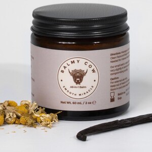 May include: A dark amber glass jar with a black screw-top lid, labeled Balmy Cow All-in-1 Balm, Net Wt. 60 mL / 2 oz. The label features a cow illustration, dried chamomile flowers, and a vanilla bean pod placed in front of the jar.