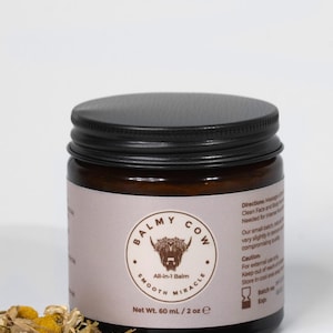 May include: A dark amber glass jar with a black screw-top lid, containing 60 mL / 2 oz of Balmy Cow All-in-1 Balm. The label features a highland cow illustration, text, and directions. Dried chamomile flowers and a vanilla bean lie in front.