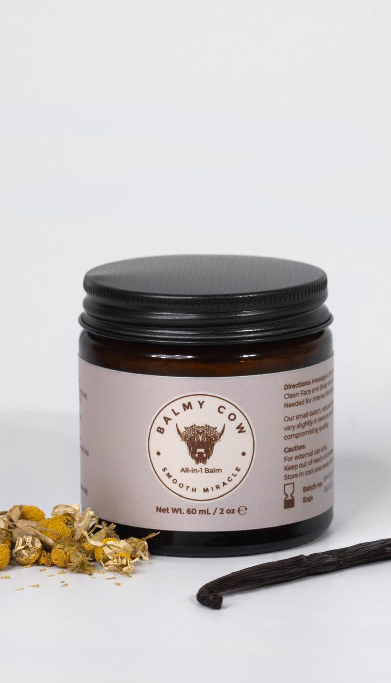 May include: A dark amber glass jar with a black screw-top lid, containing 60 mL / 2 oz of Balmy Cow All-in-1 Balm. The label features a highland cow illustration, text, and directions. Dried chamomile flowers and a vanilla bean lie in front.