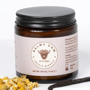 May include: A brown glass jar with a black screw-top lid containing Balmy Cow All-in-1 Balm. The label features a cow illustration, the text Net Wt. 120 mL / 4 oz, and directions. Dried chamomile flowers and a vanilla bean are placed in front.
