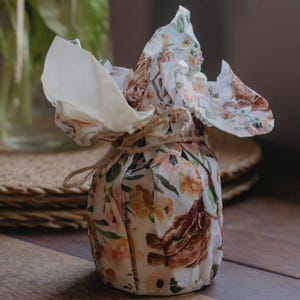 May include: A small, round object wrapped in white paper featuring a repeating pattern of brown Highland cattle and orange floral illustrations. The paper is gathered at the top and secured with a thin, natural-colored jute twine tied in a bow.