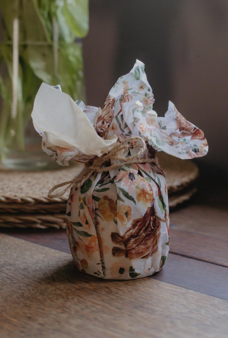 May include: A small, round object wrapped in white paper featuring a repeating pattern of brown Highland cattle and orange floral illustrations. The paper is gathered at the top and secured with a thin, natural-colored jute twine tied in a bow.