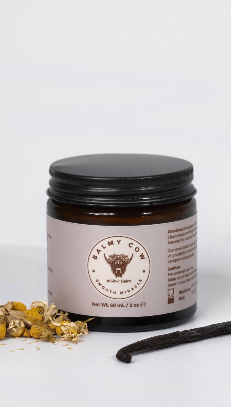 May include: A dark amber glass jar with a black screw-top lid, labeled Balmy Cow All-in-1 Balm, Net Wt. 60 mL / 2 oz. The label features a cow illustration, dried chamomile flowers, and a vanilla bean pod placed in front of the jar.