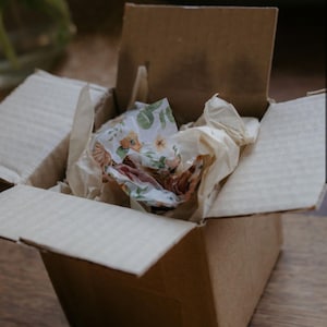 May include: An open brown cardboard shipping box containing crumpled plain beige tissue paper and a single sheet of white tissue paper decorated with a repeating pattern of green leaves and small orange flowers tucked inside.