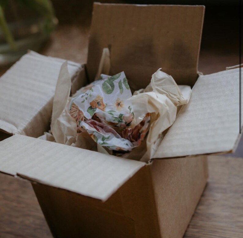 May include: An open brown cardboard shipping box containing crumpled plain beige tissue paper and a single sheet of white tissue paper decorated with a repeating pattern of green leaves and small orange flowers tucked inside.