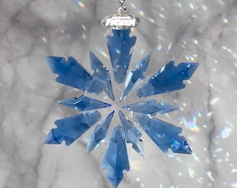 May include: A blue crystal snowflake ornament with a silver-colored metal top and a string of clear beads. The snowflake has multiple pointed edges and is suspended against a marble background.