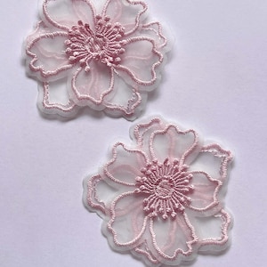 May include: Two delicate, light pink floral appliques. Each applique features a detailed embroidered outline and center, with a sheer, petal-like design. The appliques are set against a white background, showcasing their intricate details.