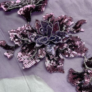 Puede incluir: Aplique floral de lentejuelas moradas con un respaldo de malla. El aplique presenta una flor grande con flores y hojas más pequeñas.