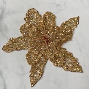 Op de afbeelding: Een gouden bloemenapplicatie met pailletten en ingewikkelde details. De bloem heeft meerdere bloemblaadjes en een centrale cluster, allemaal bedekt met glinsterende pailletten. De applicatie is ontworpen voor verfraaiing en voegt een vleugje glamour toe aan elk project.