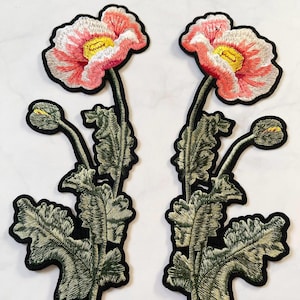 May include: Two embroidered poppy flower patches. The flowers have pink and white petals with yellow centers. The green leaves and stems are detailed with embroidery. Each patch has a black border.