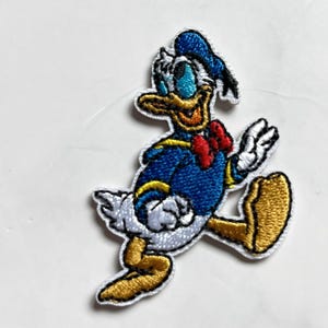Op de afbeelding: Geborduurde patch met Donald Duck in een rennende pose. De patch toont Donald Ducks blauwe matrozenhemd, hoed en rode vlinderdas. De patch is wit omlijnd en heeft goudkleurige voeten.