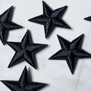 May include: Close-up of several black star-shaped embroidered patches. The patches are made of a textured fabric and are arranged on a white surface. The stars have five points and are evenly spaced. These patches are suitable for various craft projects.