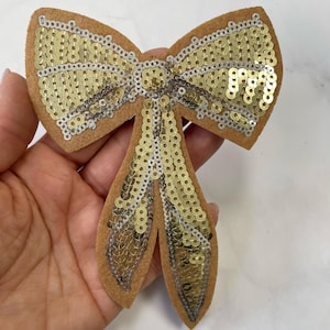 May include: A tan felt bow with gold sequins and silver trim. The bow is in a classic bow shape, with two loops and two streamers. The sequins are densely packed, creating a shimmering effect. This decorative bow is suitable for crafts and embellishments.