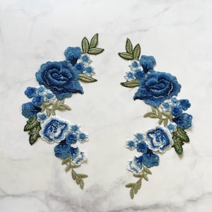 May include: Two embroidered floral patches featuring blue roses and smaller white and blue flowers with green leaves. The patches are arranged in a curved, symmetrical design, suitable for embellishing clothing or crafts.