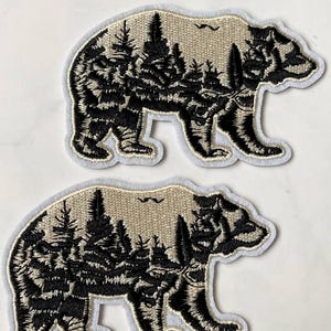 May include: Two embroidered iron-on patches featuring a black bear silhouette with a forest scene inside. The bear is outlined in white and the background is beige.