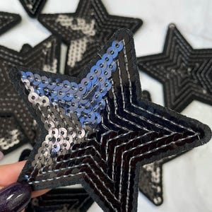 Può includere: Una toppa a forma di stella nera con un design di paillettes. Le paillettes sono nere e argentate e creano un effetto scintillante. La toppa è realizzata in feltro e ha un contorno cucito.