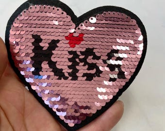 Puede incluir: Parche de lentejuelas en forma de corazón rosa con la palabra "Kiss" en lentejuelas negras. El parche tiene un respaldo de fieltro negro.
