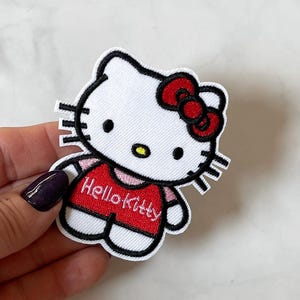 May include: Embroidered Hello Kitty patch. The patch shows a white head with a red bow, black eyes, and a yellow nose. She wears a red shirt with the words "Hello Kitty" in white.