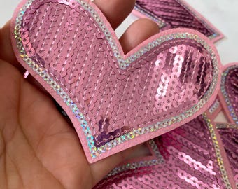 Puede incluir: Paillettes rosas en forma de corazón con un borde blanco. Las lentejuelas están cosidas sobre un soporte de fieltro rosa.