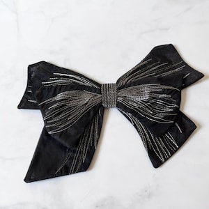 May include: A large, black bow with silver thread detailing. The bow features a textured center and long, flowing ends. The silver thread creates a striking contrast against the dark fabric, adding a touch of elegance. This decorative bow is suitable for various craft projects.