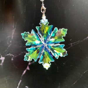May include: A decorative snowflake ornament with iridescent green and blue crystal facets. The snowflake is suspended by a silver-colored clasp and a string of clear beads. The ornament is designed to catch and reflect light.