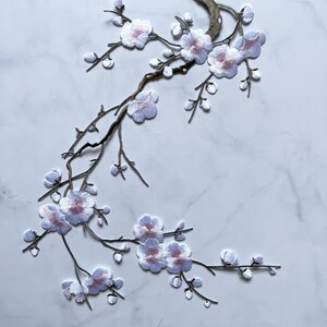 Puede incluir: Parche bordado de rama floral con flores blancas y rosas. El parche tiene un tallo marrón y está diseñado para coserlo a la tela. Las flores tienen un aspecto delicado y en capas, sobre un fondo de mármol blanco.