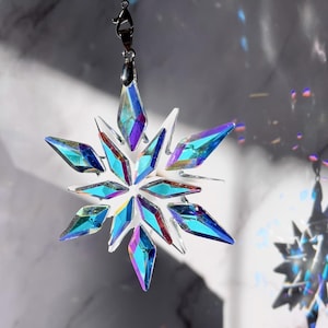 Puede incluir: Adorno colgante de copo de nieve de cristal iridiscente. El adorno está hecho de múltiples piezas de cristal claras y facetadas que reflejan la luz azul, púrpura y verde. Se suspende mediante un cierre de metal plateado y una cuerda transparente.