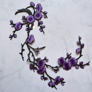 May include: Embroidered floral applique with purple flowers and buds on black stems. The design is a stylized flowering branch, suitable for fabric embellishment or craft projects.