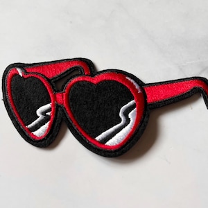 May include: Embroidered patch of heart-shaped sunglasses. The sunglasses have black lenses, a red frame, and white and black detailing. The patch is designed to be ironed or sewn onto fabric, suitable for clothing or accessories.